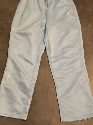 Nike NOCTA ljusblå mjukisbyxor - Säljer ett par ljusblå mjukisbyxor från Nike NOCTA-kollektionen. Byxorna har bred passform, dragsko i midjan och dragkedjeförsedd ficka. Swoosh-logga på höger sida och NOCTA-tryck på vänster ben. Perfekta för chill dagar och streetwear.