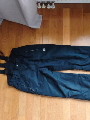 Svarta täckbyxor från 157 Funk - Säljer ett par svarta täckbyxor från 157 Funk med justerbara hängslen och dragkedjor vid fickorna. Byxorna har hög midja och quiltat foder för extra värme. Perfekta för kalla vinterdagar och snöaktiviteter.