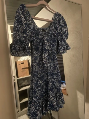 Blå blommig offshoulder klänning - Söt blå kortklänning med vita blommor, offshoulder-modell och puffiga kortärmar. Klänningen har volangkant nertill och en dekorativ knytning framtill. Perfekt för sommarens alla tillfällen.
