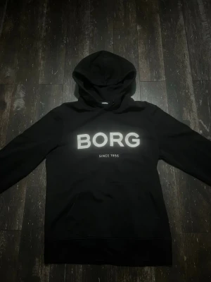 Svart hoodie från BORG - Cool svart hoodie från BORG med stor vit logotyp på bröstet och texten 'SINCE 1956' under. Klassisk huva och stor framficka. Perfekt för dig som gillar sportig och avslappnad stil. Materialet är mjukt och bekvämt, troligen bomullsblandning. Hör av er vid frågor 