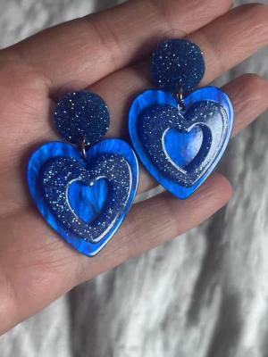 Blå hjärtformade örhängen - Snygga örhängen i form av hjärtan med intensiv blå färg och glittrig yta. De är tillverkade i plast eller akryl och har en modern, lekfull design som verkligen sticker ut. Perfekta för dig som vill addera lite färg och glans till din stil.