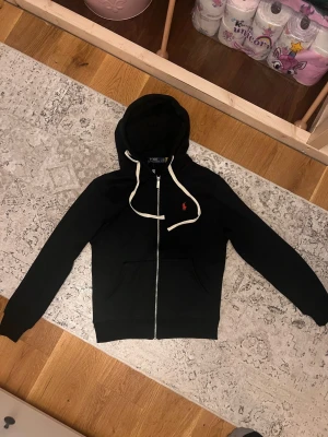 Svart hoodie från Polo Ralph Lauren - Svart hoodie med dragkedja från Polo Ralph Lauren. Klassisk design med vit dragsko i huvan och röd broderad logga på bröstet. Mjuk bomullskvalitet och två fickor framtill. Perfekt för en avslappnad och stilren look.