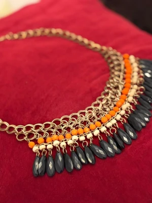 Kedjehalsband med färgglada pärlor - Unikt halsband med grov guldfärgad kedja och dekorativa pärlor i orange, beige och svarta droppformade hängen. Halsbandet har flera rader av kedjor och pärlor som ger ett statement-utseende. Perfekt för dig som vill sticka ut med accessoarer.