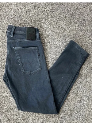 Mörkgrå slim fit jeans från G-Star - Snygga mörkgrå jeans replay med slim fit-modell och klassiska fem fickor. Jeansen har en stilren design med svarta detaljer och replay -logga på bakfickan. Perfekta för en trendig och avslappnad look.