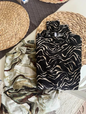 Två mönstrade polotoppar  - Säljer två snygga polotoppar från Hays i storlek M. Ena toppen är svart med vitt/beige zebramönster och den andra har ett mönster i beige, brunt och grönt. Båda är väldigt mjuka och håller en varm på vår/höst/vinter. Passar även L samt XL skulle jag säga!