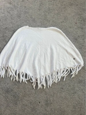 Vit sjal med fransar och hjärtdetalj - Säljer en vit sjal med långa fransar längs kanten och en liten metallhjärta som detalj. Sjalen har en mjuk och flowy känsla, perfekt att slänga över axlarna för en chill look. Materialet känns som bomull eller viskos.