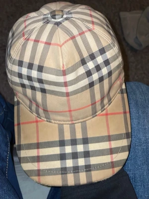 Beige keps från Burberry med rutmönster - Säljer en klassisk beige keps från Burberry med det ikoniska rutmönstret i svart, vitt och rött. Kepsen har böjd skärm och justerbar passform. Tillverkad i bomull för en bekväm känsla och snygg look. Perfekt accessoar för att lyfta din outfit.