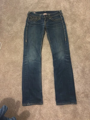 Mörkblå bootcut jeans från True Religion - Säljer ett par mörkblå bootcut jeans från True Religion med kontrastsömmar och klassiska fem fickor. Jeansen har en snygg tvätt och detaljerad ficka fram med knapp. Perfekta för dig som gillar en avslappnad men trendig stil. Pris kan diskuteras !!