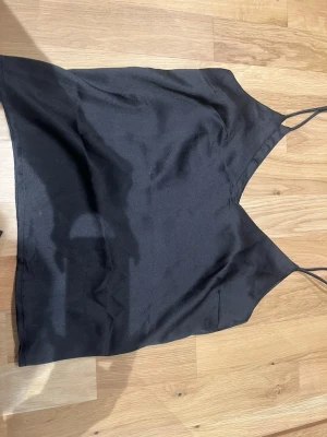 Svart linnetopp från The Black Label - Snygg svart linnetopp från The Black Label Collection i storlek XS. Toppen har tunna axelband och v-ringad halsringning, perfekt för en stilren och enkel look. Materialet har en lätt glansig finish som ger en exklusiv känsla.