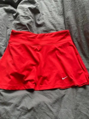 Röd tenniskjol från Nike - Röd sportkjol från Nike i stretchigt funktionsmaterial. Perfekt för tennis, padel eller annan träning. Ser inte vart det står storlek men jag har S i allt och den passar perfekt 