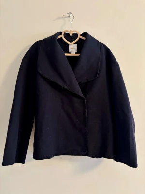 Marinblå ullkappa med bred krage - Stilren marinblå kappa i ull med bred, markerad krage och rak passform. Kappan har minimalistisk design utan synliga knappar och är något kortare i modellen. Perfekt för dig som gillar en clean och modern look.