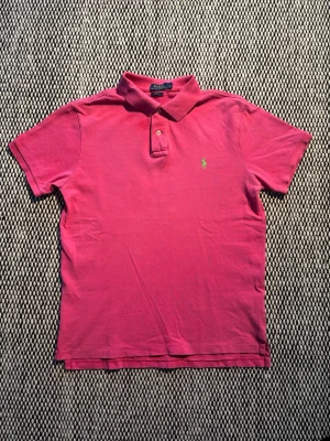 Rosa pikétröja från Ralph Lauren - Klassisk rosa pikétröja från Ralph Lauren med krage och två knappar framtill. Tröjan har det ikoniska gröna broderade logotypen på bröstet och är tillverkad i bomull. Perfekt för en avslappnad men ändå stilren look.