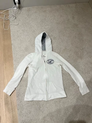 Vit zip hoodie från Hollister - Vit hoodie från Hollister med dragkedja framtill och klassisk logga på bröstet. Tillverkad i mjuk bomullsblandning (60% bomull, 40% polyester). Har kängurufickor och huva med grått innerfoder. Perfekt för en avslappnad och stilren look.