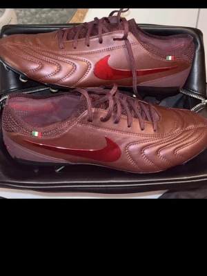 Exklusiva Nike Tiempo Legend fotbollsskor brun - Snygga Nike Tiempo Legend fotbollsskor som ändast finns i ett särskilt antal, ca 2000.   I metallicbrunt skinn med röda detaljer och klassisk Swoosh på sidan. Skorna har italiensk flaggdetalj vid hälen och är designade för gräsplan. Levereras i exklusiv svart väska med 'Legend Tiempo' tryck. 
