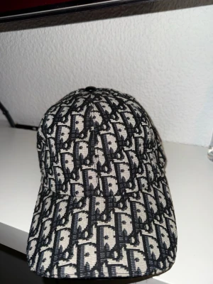 Svartvit keps från Dior med monogram - Säljer en snygg svartvit keps från Dior med det klassiska Dior-monogrammet över hela kepsen. Kepsen har böjd skärm och justerbart spänne bak. Tillverkad i ett slitstarkt tyg med exklusiv känsla och detaljerad insida med Dior-logga.