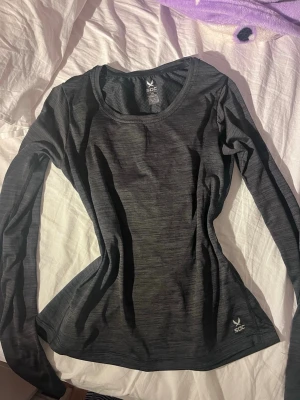 Mörkgrå långärmad träningströja SOC - Snygg mörkgrå långärmad träningströja från SOC i tekniskt funktionsmaterial. Tröjan har rund halsringning och diskret melerad design, perfekt för löpning eller gym. Materialet är lätt och andas, vilket gör den skön under intensiva pass.