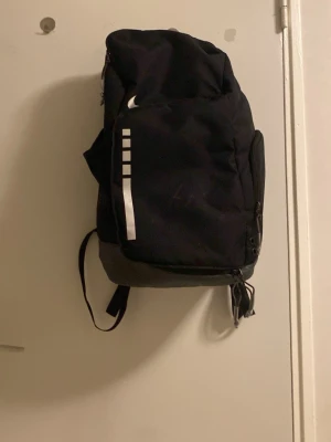 Svart ryggsäck från Nike - Snygg svart ryggsäck från Nike med vita detaljer och logga. Ryggsäcken har vadderade axelband med reflekterande partier och flera fack med dragkedja. Perfekt för skola eller träning och rymmer allt du behöver. Tillverkad i slitstarkt material.