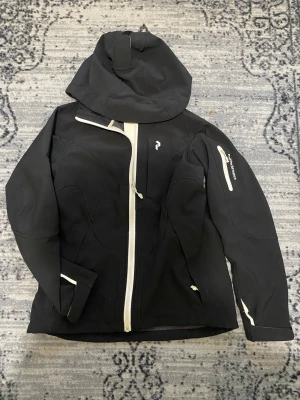 Svart vindjacka från Peak Performance - Snygg svart vindjacka från Peak Performance med vit dragkedja och detaljer. Jackan har huva, ficka på ärmen med dragkedja och logga på bröstet. Tillverkad i ett mjukt och smidigt material som passar perfekt för blåsiga dagar.