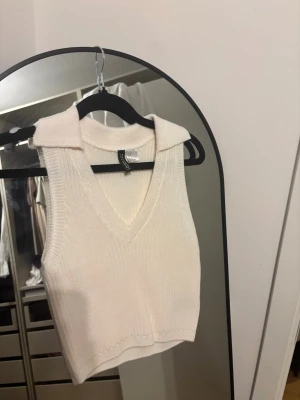 Vit ribbad stickad linnetopp H&M - Säljer en vit ribbad stickad linnetopp från H&M Divided. Toppen är ärmlös, croppad och har en snygg krage samt v-ringning. Perfekt att styla med jeans eller kjol för en trendig look.