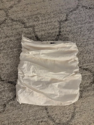 Vit omlott bandeau topp från Zara - Säljer en vit bandeau tubtopp med snygga rynkade detaljer från Zara. Toppen är axelbandslös! Den bruna detaljen där bak på tyget går att stoppa in bakom tröjan om man inte vill att den syns 💕