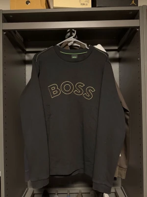 Svart sweatshirt från BOSS - Stilren svart sweatshirt från BOSS med stor logga i guld på bröstet. Tröjan har rund halsringning, långa ärmar och ribbade muddar vid ärmslut och nederkant. Perfekt för en avslappnad och trendig look.