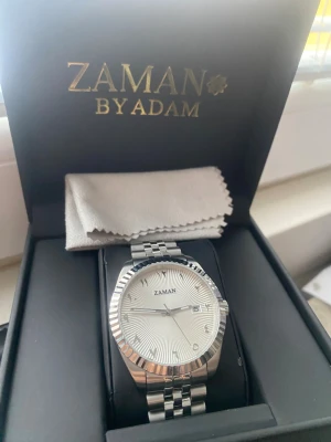 Zaman By Adam silverfärgad klocka - Stilren klocka från Zaman By Adam med silverfärgat armband i metall och vit urtavla med subtilt mönster. Klockan har datumvisning och klassisk länkdesign. Perfekt accessoar för dig som gillar en clean och modern look.