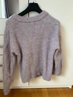 Ljuslila kabelstickad tröja - Mysig ljuslila stickad tröja med kabelstickat mönster och bred ribbad halsringning. Tröjan har långa ärmar och ribbade muddar vid ärmslut och nederkant. Perfekt för att hålla sig varm under kyliga dagar.