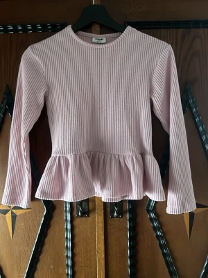 Rosa ribbad topp med volangkant - Supersöt ljusrosa topp från Kappahl med ribbad struktur och volangkant nedtill. Långärmad modell med rund halsringning, perfekt för dig som gillar en feminin och trendig stil. Passar till både jeans och kjol för en chill look.