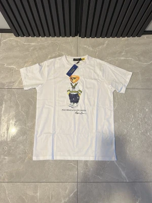 Vit Polo Bear t-shirt från Ralph Lauren - Vit t-shirt från Polo Ralph Lauren med ikoniskt Polo Bear-tryck på bröstet. Klassisk passform med rund hals och korta ärmar. Tillverkad i mjuk bomull, perfekt för en avslappnad och stilren look.