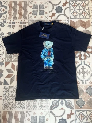 Marinblå t-shirt Polo Ralph Lauren - Snygg marinblå t-shirt från Polo Ralph Lauren med ikoniska björnmotivet på bröstet. Klassisk rund hals och korta ärmar. Tillverkad i mjuk bomull som känns skön mot huden. Perfekt för dig som vill ha en stilren men lekfull look.