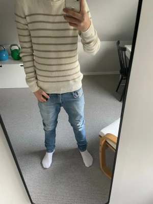 Beige och vit randig stickad tröja - Snygg stickad tröja i beige och vitt med horisontella ränder från J Lindberg . Tröjan har rund halsringning, långa ärmar och ribbade muddar vid ärmslut och nederkant. Perfekt för en avslappnad och trendig look.