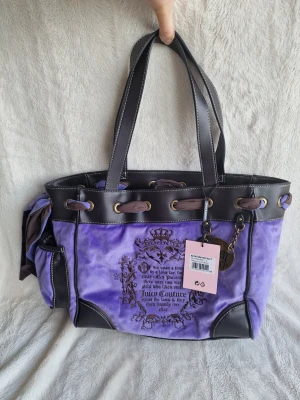 Juicy Couture daydreamer fairytale tote handväska i velour - Superfin lila handväska från Juicy Couture i mjuk velour med bruna detaljer i konstläder. Väskan har tryckt logga och text framtill, dekorativ rosett på sidan och metallberlocker. Två långa handtag och snygga guldfärgade metalldetaljer.
