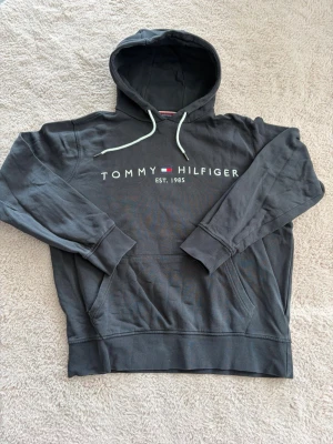Mörkgrå hoodie från Tommy Hilfiger - Snygg mörkgrå hoodie från Tommy Hilfiger med vit dragsko och stor logga framtill. Tillverkad i ekologisk bomull och har en klassisk känguruficka. Perfekt för en avslappnad och trendig stil. Står L men den sitter som en S