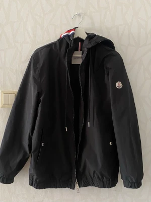Svart Moncler jacka  - Svart vindjacka från Moncler med huva och dragkedja. Jackan har två fickor med knappar och Moncler-logga på vänster ärm. Huva med röd, vit och blå detalj. Materialet är lätt och vattenavvisande, perfekt för blåsiga dagar.