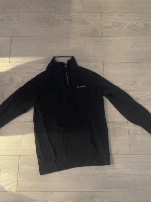 Svart halvzip-tröja från Bondelid - Svart långärmad tröja från Bondelid med halv dragkedja och liten broderad logga på bröstet. Tröjan har ribbade muddar vid ärmslut och nederkant, perfekt för en stilren och avslappnad look.