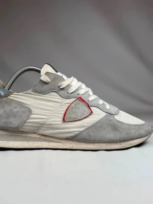 Philippe Model  - Snygga sneakers från Philippe Model i grått och vitt med mockadetaljer och mesh. Skorna har en ikonisk sköldlogga med röd kant på sidan och vita skosnören. Låg modell med bekväm sula och svart detalj på plösen. Skorna är storlek 43 men passar 44. 