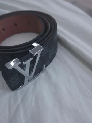  Damier svart bälte - Snyggt svart bälte från med klassiskt Damier-mönster och en stor silverfärgad  som spänne. Bältet är gjort i läder och har en känsla som lyfter vilken outfit som helst.