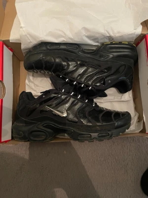 Nike Air Max Plus svarta sneakers - Svarta Nike Air Max Plus sneakers med silvrig Swoosh-logga på sidan. Skorna har en vågig design i syntetmaterial och mesh, samt svarta snören och en robust sula med synliga luftbubblor. Perfekta för dig som gillar en sportig och edgy stil.