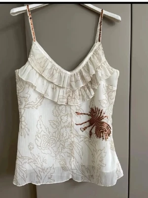 Vit linnetopp med volang och paljetter - Supersöt vit linnetopp med tunna axelband, volangdetalj framtill och diskret blommigt mönster. Toppen har en v-ringning och en dekorativ paljettapplikation i kopparfärg på ena sidan. Perfekt för sommarens alla tillfällen!