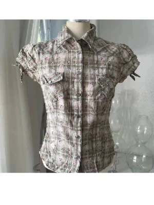 Rutig kortärmad skjorta med blommor - Kortärmad skjorta med rutigt mönster i beige och grönt , dekorerad med små rosa blommor. Skjortan har knappar framtill, två bröstfickor och snörning vid ärmsluten för en cool detalj. Perfekt för dig som gillar en söt men avslappnad stil.