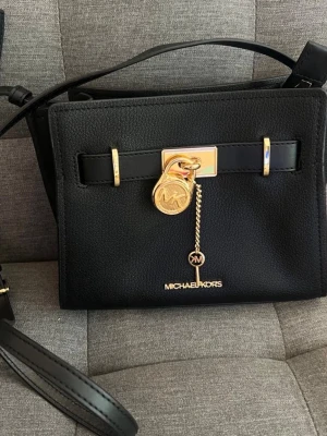 Svart handväska från Michael Kors - Snygg svart handväska från Michael Kors med guldfärgade detaljer och dekorativt hänglås framtill. Väskan har justerbar axelrem. Perfekt för dig som vill ha en stilren och trendig accessoar.