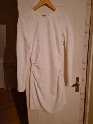 Vit långärmad klänning från Kappahl - Säljer en vit långärmad klänning från NAKD med strukturerat tyg och rund halsringning. Klänningen är kort och har en figurnära passform som framhäver siluetten. Endast använd en gång