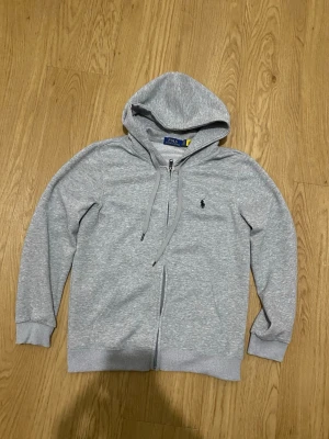 Grå zip hoodie från Polo Ralph Lauren - Snygg ljusgrå hoodie med dragkedja från Polo Ralph Lauren. Klassisk design med huva och dragsnören, ribbade muddar och det ikoniska lilla Polo-logotrycket på bröstet. Tillverkad i mjuk bomullsblandning som är perfekt för chill dagar.
