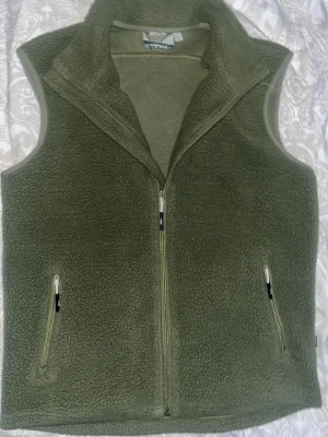 Olivgrön fleeceväst från Tuxer - Mysig olivgrön fleeceväst från Tuxer med hög krage och hel dragkedja framtill. Västen har två praktiska fickor med dragkedja och broderad logga på ryggen. Perfekt lager för kyliga dagar och outdoor-äventyr.