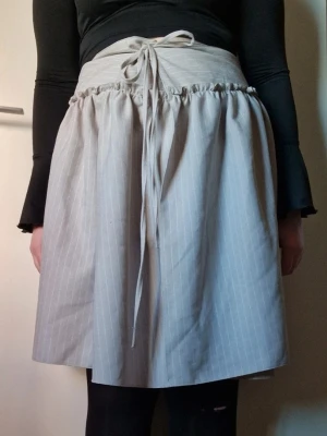 Midikjol med snörning - Snygg beige kjol med diskreta vita ränder och hög midja. Kjolen har en bred linning med snörning framtill som ger en cool och unik look. Modellen är A-linjeformad och tyget känns mjukt och bekvämt, perfekt för dig som gillar stilrena plagg med twist. Den har en knapp på sidan och sen knyts den i midjan. Slutar vid knäna på mig ( jag är 168 cm lång ). Något nopprig och någon lös tråd finns men på hela taget är den i fint skick. Storlek 40. Katt finns i hemmet. Jag har fler klädannonser uppe