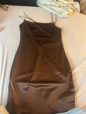 Brun satin klänning med smala band - Snygg brun kort klänning i satin med smala axelband och öppen rygg med snörningsdetalj. Klänningen har en figurnära passform och glansigt material som ger en lyxig känsla. Perfekt för dig som vill sticka ut med en trendig look.