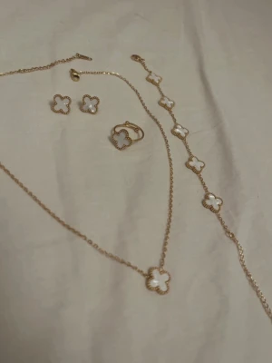 Guldpläterat smyckeset med fyrklöver - Snyggt smyckeset med halsband, armband, ring och örhängen i guldplätering. Alla delar har vita fyrklöverformade hängen med pärlliknande kant. Perfekt för dig som gillar eleganta och trendiga accessoarer.