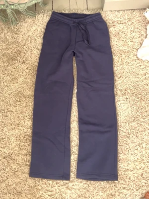 Oanvända Mörkblå mjukisbyxor - Mörkblå mjukisbyxor med raka ben och resår i midjan samt dragsko för justerbar passform. Tillverkade i mjukt och bekvämt material, perfekta för en avslappnad stil. Inga synliga loggor eller märken.