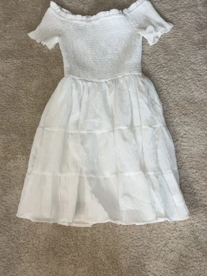Vit offshoulder sommarklänning - Söt vit klänning med offshoulder och korta ärmar. Överdelen är smockad och kjolen har volanger för en luftig känsla. Perfekt för varma dagar och har en romantisk vibe.