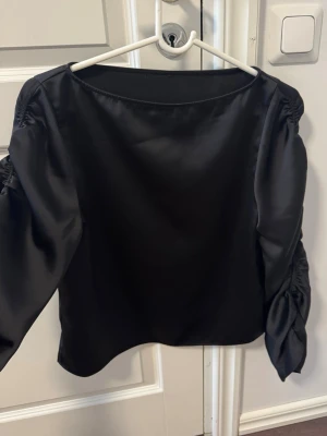 Svart satinblus med puffärmar - Stilren svart blus i glansigt satinmaterial med långa puffärmar och rynkade detaljer längs ärmarna. Blusen har en rak passform och rund halsringning. 